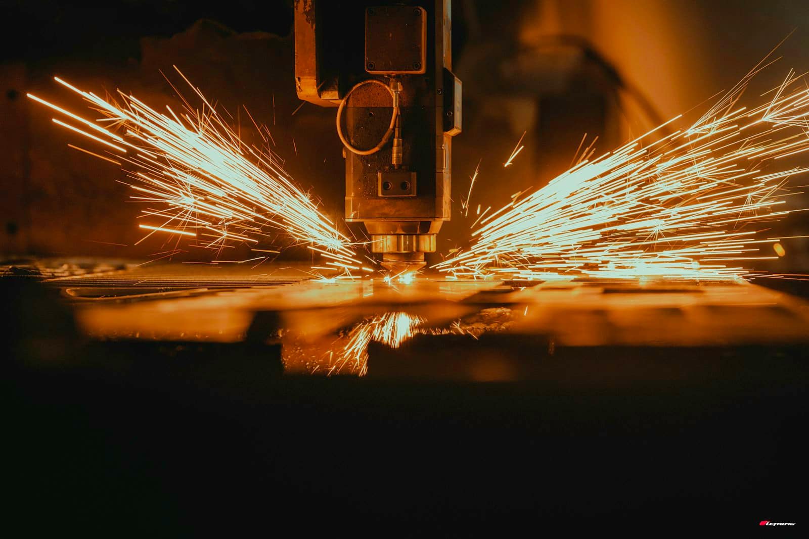 A laser cutting head throwing fans of sparks across a steel sheet in a dark workshop